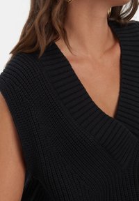 Haut noir sans manches en maille avec un col en V profond, une texture côtelée et des détails subtils le long du col et des ouvertures des bras.