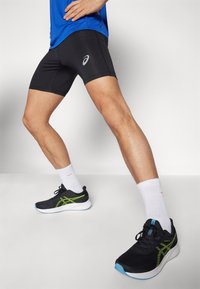 Svarta atletiska shorts med en logotyp, svarta och gröna löparskor samt vita strumpor, visade på en modell i stående position.