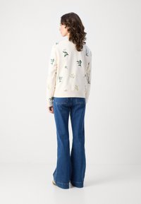 Kremfarget hettegenser med blomsterbroderi, kombinert med flagrende denimjeans. Designet har forskjellige grønne og gule plante motiver.