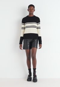 Maglione a coste in bianco e nero con righe orizzontali, abbinato a pantaloni corti in pelle nera e scarpe platform chunky, in piedi su uno sfondo bianco.