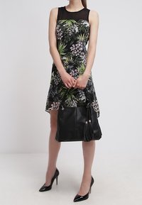 Černé květinové šaty s zelenými a růžovými vzory, bez rukávů, doplněné černou koženou taškou s třásní a stříbrným kováním.