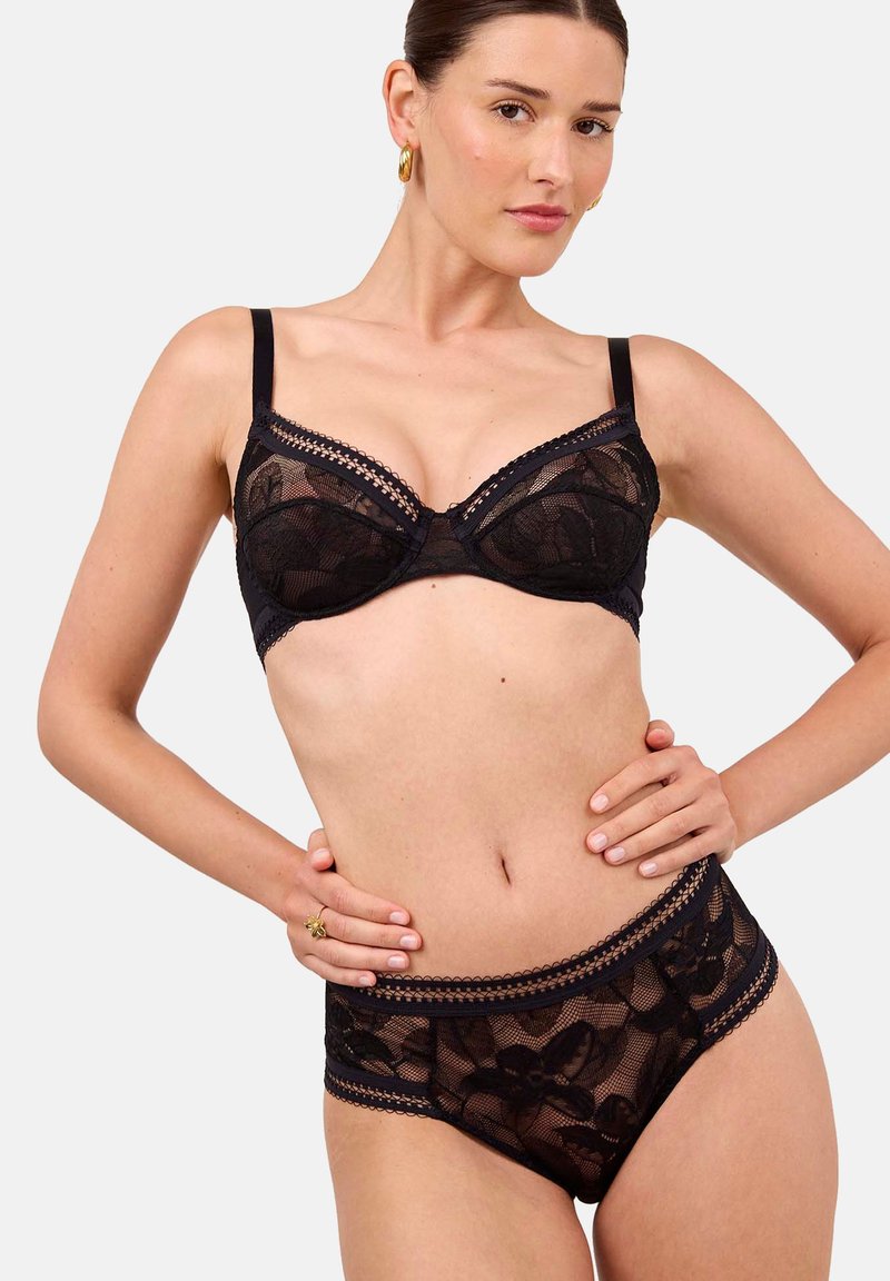 Mujer vistiendo un sujetador de encaje negro y ropa interior de encaje a juego de talle alto, posando con las manos en las caderas sobre un fondo liso.