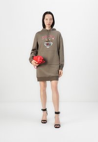 Rochie cu glugă verde măsliniu, cu grafică de tigru, buzunar frontal și tiv texturat. Ținând o geantă mică roșie. Pantofi cu toc înalt cu curea la glezna de culoare neagră.