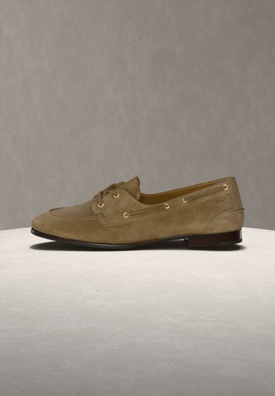 Brown suede men's loafer with lace-up front and gold eyelets, displayed on a round beige surface against a neutral background.