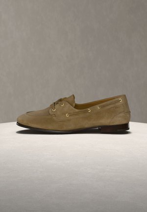 Brun herreloafer i ruskind med snørebånd foran og guld-øjer, vist på en rund beige overflade mod en neutral baggrund.