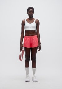 Γυναίκα όρθια, φοράει λευκό αθλητικό σουτιέν, κοραλλί σορτς Under Armour, λευκές κάλτσες και αθλητικά παπούτσια, κρατώντας ροζ λάστιχο αντίστασης στο αριστερό χέρι.