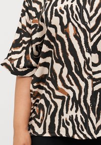 Chemise à boutons avec un imprimé zèbre noir, brun et crème, avec des manches retroussées et de petits boutons noirs le long de la patte de boutonnage.