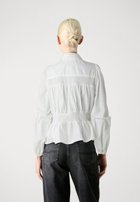 Bluz�ă albă cu talie peplum și detalii plisate la spate, având mâneci lungi și guler clasic. Asortată cu blugi negri din denim.