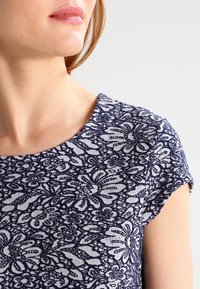 Gros plan sur une personne portant un haut à manches courtes bleu marine à motifs floraux blancs, montrant le cou, l'épaule et les lèvres.