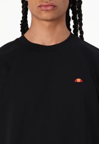 Black cotton t-shirt with a rounded neck, featuring a small embroidered logo in orange and red on the left chest area.
