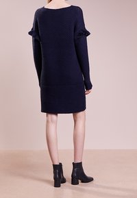 Abito maglione blu navy con texture a coste, maniche lunghe e dettagli a balze sulle spalle. Abbinato a stivaletti neri alla caviglia.