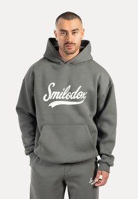 Szara bluza z kapturem z przednią kieszenią i białym logo "Smilodox". Miękka tekstura i luźny krój, w połączeniu z dopasowanymi szarymi spodniami.