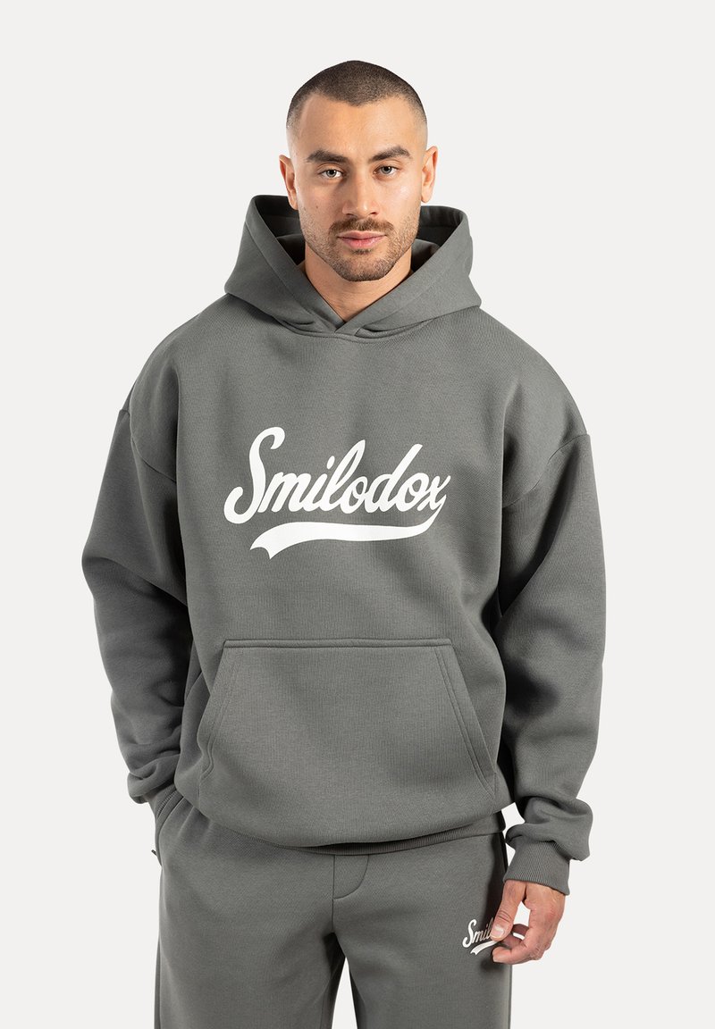 Szara bluza z kapturem z przednią kieszenią i białym logo "Smilodox". Miękka tekstura i luźny krój, w połączeniu z dopasowanymi szarymi spodniami.