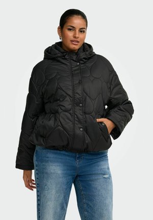 Frau trägt eine schwarze gesteppte Jacke mit Herzmustern und blauer Jeans, steht mit einer Hand in der Tasche vor einem schlichten Hintergrund.