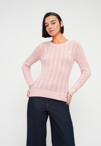 Femeie îmbrăcată într-un pulover roz deschis cu model torsadat și blugi evazați bleu închis, stând cu mâinile pe șolduri, pe un fundal simplu și luminos.