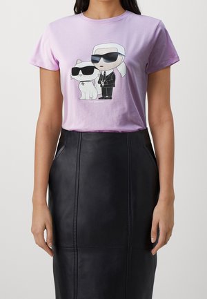 Lilan värinen t-paita, jossa on sarjakuvakuva kissasta ja aurinkolaseja käyttävästä henkilöstä. Henkilöllä on valkoiset hiukset ja puku. Musta nahkahame.