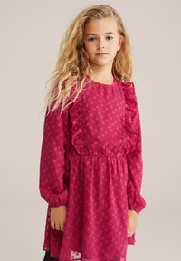 Mlada deklica z dolgimi valovitimi blond lasmi, oblečena v teksturirano fuksijasto obleko z volančki in dolgimi rokavi, proti enotni ozadju.