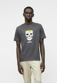 Šedé bavlnené tričko s veľkým grafickým motívom lebky so žltým viečkom, umiestneným centrálně, skombinované s béžovými nohavicami.