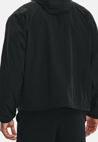 Giacca leggera nera con cappuccio, caratterizzata da una texture liscia e una vestibilità rilassata. Maniche lunghe e orlo posteriore allungato sono visibili.