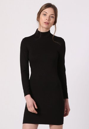 Joven con cabello castaño claro que lleva un vestido negro ajustado de manga larga y cuello alto, de pie contra un fondo claro y sencillo.