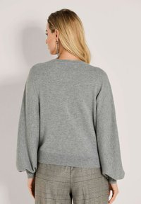 Frau mit langen blonden Haaren, die einen grauen Pullover mit Ballonärmeln und karierter Hose trägt, mit dem Rücken zu einer einfarbigen Hintergrund. 