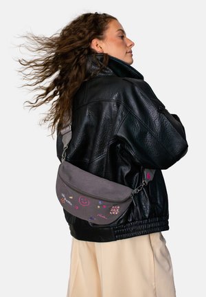 Young woman with wavy hair wearing a black leather jacket and beige pants, carrying a grey crossbody bag with colorful doodles.