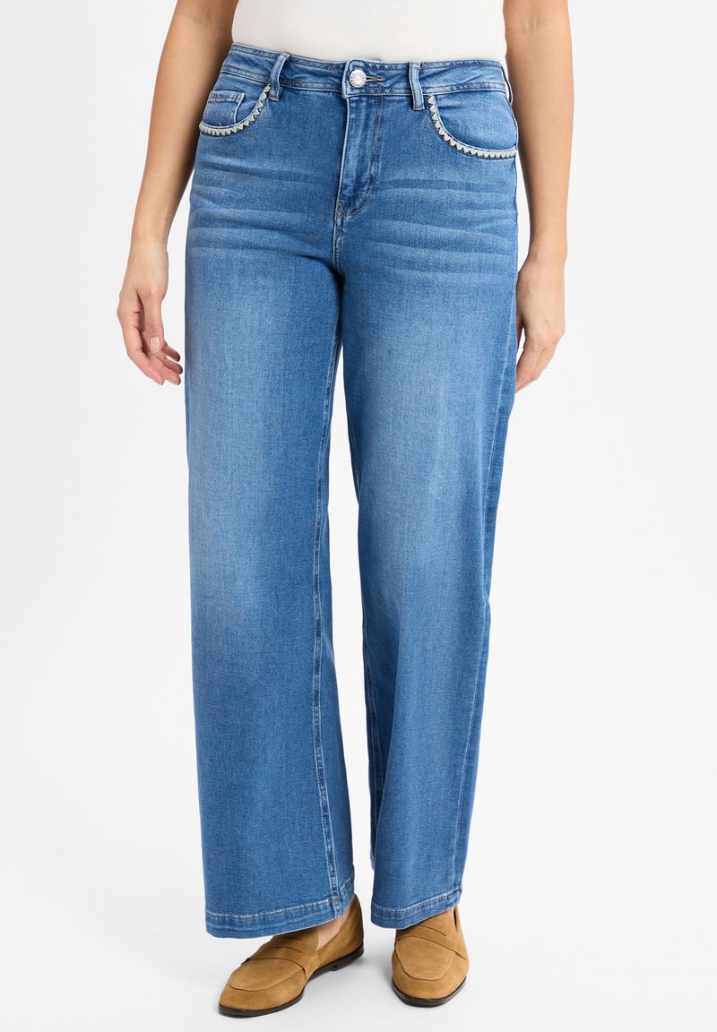 Blauwe denim wijde jeans met subtiele vervaging, vijf zakken en decoratieve stiksels langs de voorkant van de zakken, gecombineerd met bruine instappers.