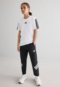 Weißes Adidas-T-Shirt mit schwarzen Streifen an den Ärmeln; schwarze Hose mit weißen Streifen und Logo; getragen mit weißen Sportschuhen.