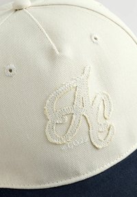 Többnyire bézs és tengerészkék baseball sapka, közelről, a homlokrészén textúrázott, varrott "Ag" logóval.