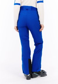 Blauwe skibroeken met een taps toelopende snit, voorzien van een gestructureerde tailleband en zwarte accenten bij de enkel. Gdragen met zwarte skibotten.
