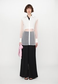 Sheer white long-sleeve shirt with a pointed collar, layered over a dark top, paired with wide black pants, and holding a pink cloth accessory.