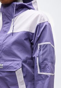 Άτομο που φοράει ένα μπουφάν ανοιχτό μοβ και λευκό με μια τσέπη από δίχτυ με φερμουάρ στο πάνω μέρος του μανικιού και μια εμπρόσθια τσέπη με καπάκι.