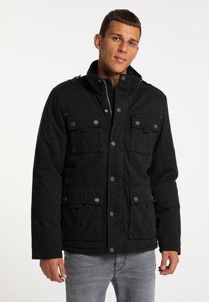 Jeune homme portant une veste noire boutonnée avec plusieurs poches avant et un jean gris clair sur un fond clair uni.