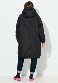 Abrigo negro aislante con capucha, de corte largo, con elástico en el dobladillo y bolsillos laterales. Se lleva con jeans oscuros y botas de plataforma rosas.