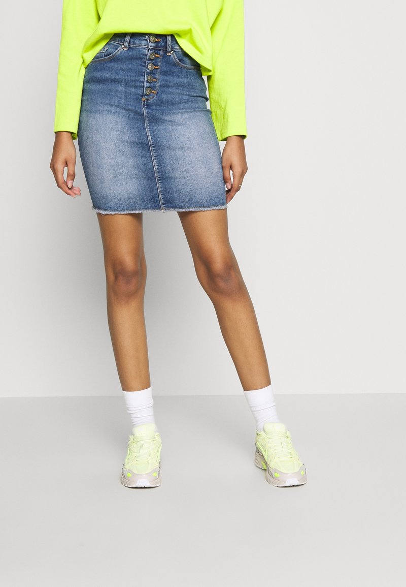 Personne portant une jupe en jean bleue à boutons sur le devant, un haut à manches longues vert néon, des chaussettes blanches et des baskets vert clair, debout sur un sol gris uni.
