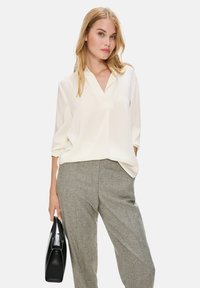 Weißes Langarmbluse mit V-Ausschnitt und lässiger Passform, kombiniert mit grauen gemusterten Hosen. Das Modell hält eine schwarze Handtasche.