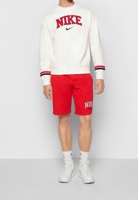 Vit sweatshirt med röd "NIKE" text och svart logotyp, parat med röda shorts med "NIKE" brodyr, bärs med vita sneakers.