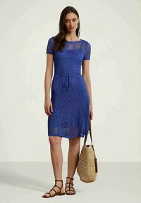 Ženska v modri kratkosleečni pleteni obleki drži tkano torbo, obuta v rjave sandale s trakovi, stoji ob ozadju v enotni barvi.