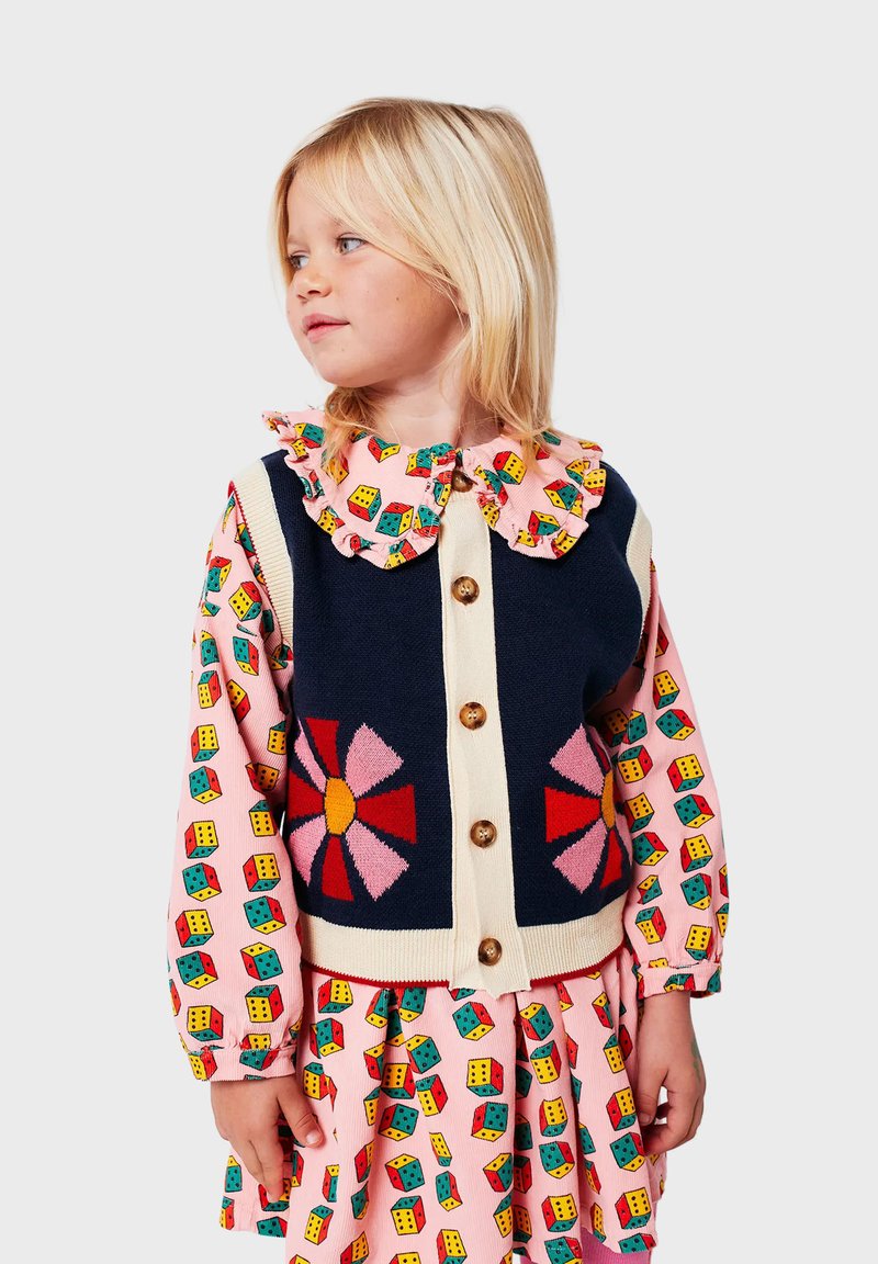 Chaleco de punto en color marino con un motivo de flores coloridas, borde crema y botones de madera, superpuesto a un vestido rosa con un patrón de dados.