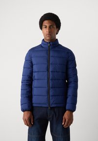 Dunkelblaue Steppjacke mit hohem Kragen, Reißverschluss und gestepptem Design. Mit elastischen Bündchen und einem Logo-Aufnäher am Ärmel.