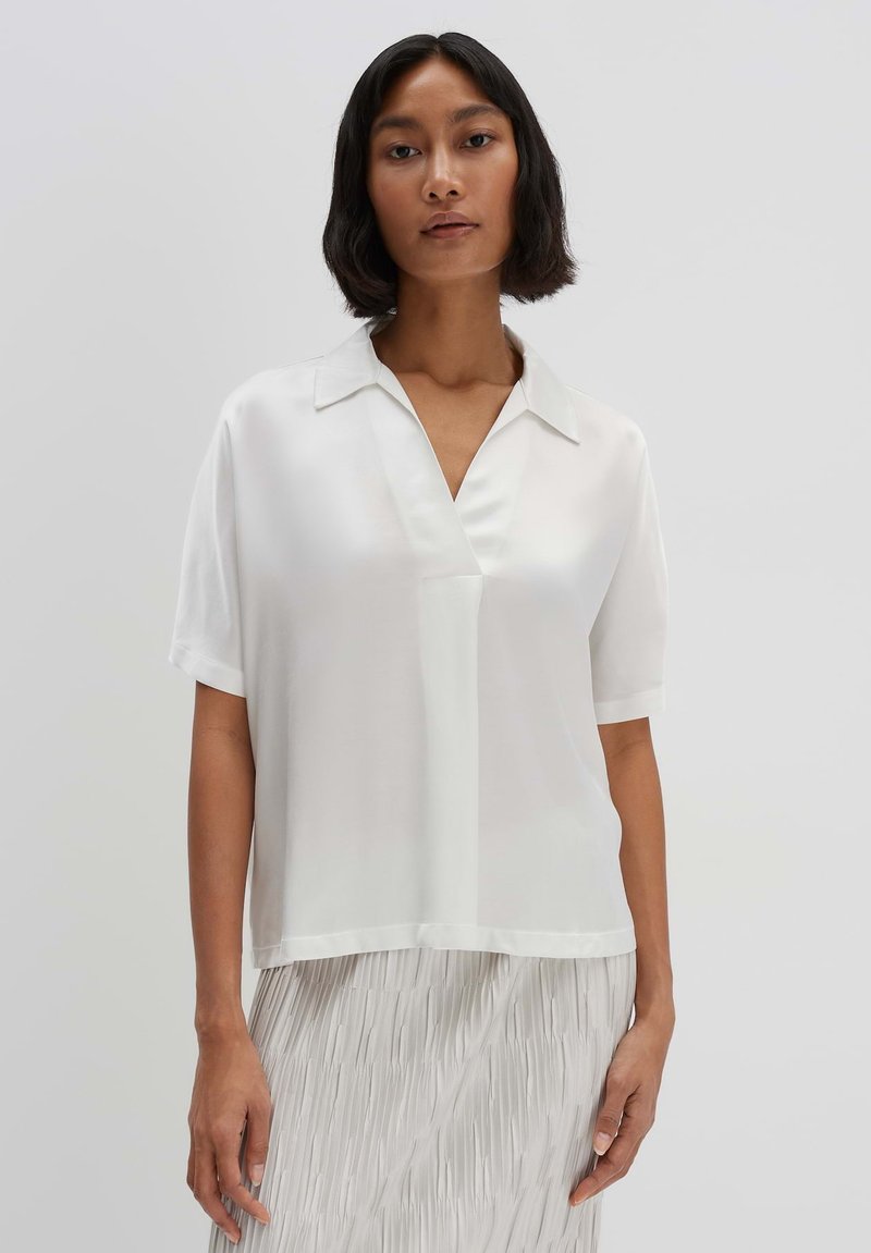 Blusa bianca a maniche corte con scollatura a V e colletto, realizzata in tessuto morbido. Presenta una vestibilità rilassata e una leggera lucentezza per un aspetto curato.