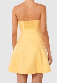 Žena v bezramínkových, světle žlutých šatech ve stylu A, zobrazená ze zadní strany na jednobarevném světle šedém pozadí.