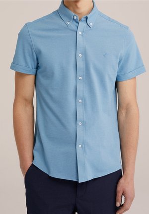 Mann trägt hellblaues kurzärmeliges Hemd mit hochgekrempelten Ärmeln und dunkle Hose, steht mit den Händen in den Taschen.