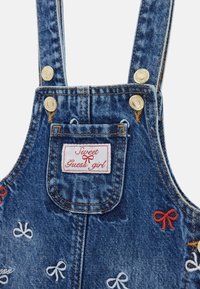 Dżinsowe ogrodniczki z regulowanymi ramiączkami, z białą etykietą z czerwonym napisem. Haftowane kokardki w kolorach czerwonym, białym i niebieskim na wyblakłej niebieskiej tkaninie.