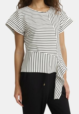 Frau trägt eine kurzärmelige weiße Bluse mit schwarzen vertikalen und horizontalen Streifen und asymmetrischem Rüschen-Detail, kombiniert mit schwarzen Hosen.