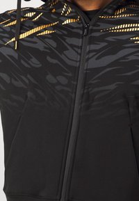 Haut zipé noir avec un motif textur é dans des nuances de gris foncé et des accents dorés. Comprend une poche frontale et une capuche doublée.