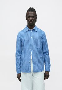 Mężczyzna o ciemnej karnacji i zaplecionych włosach, ubrany w niebieską koszulę w paski z guzikami na białą koszulkę i jasne spodnie na jednolitym tle.