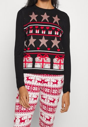 Kvinde iført sort feriesweater med stjerner, hjerter og gaveæsker, kombineret med hvide leggings med røde rensdyr- og snefnugsmønstre.