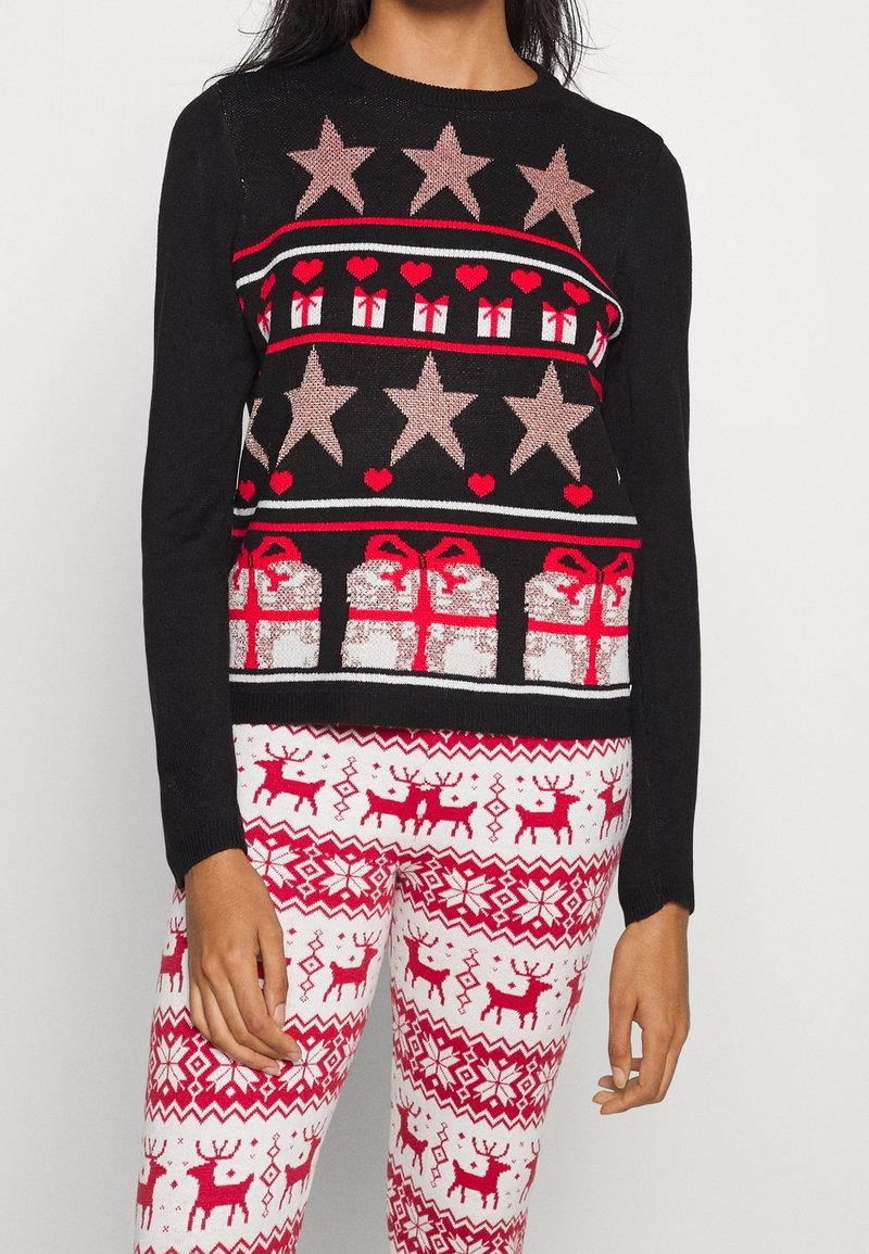 Femme portant un pull de fête noir orné d'étoiles, de cœurs et de boîtes cadeaux, associé à des leggings blancs avec des motifs de rennes rouges et de flocons de neige.