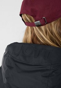 Zwarte jas met reliëf "JOTT" logo op de achterkant, gecombineerd met een bordeauxrode pet met een metalen verstelring en textielstof.